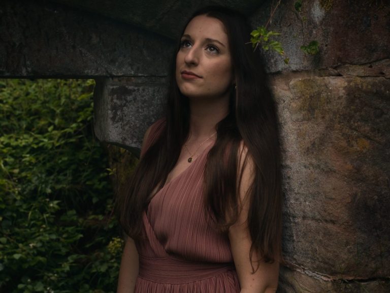 Frau in schwarzem Kleid steht lächelnd vor blühenden Kirschbäumen.