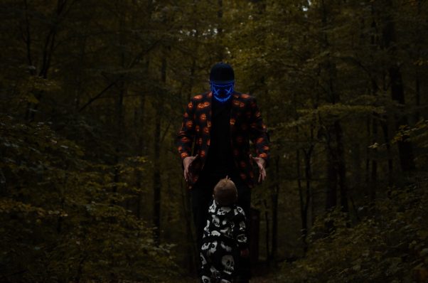 Eine Person in einem bunten Anzug steht auf einem Waldweg und blickt zu einem Kind hinunter.