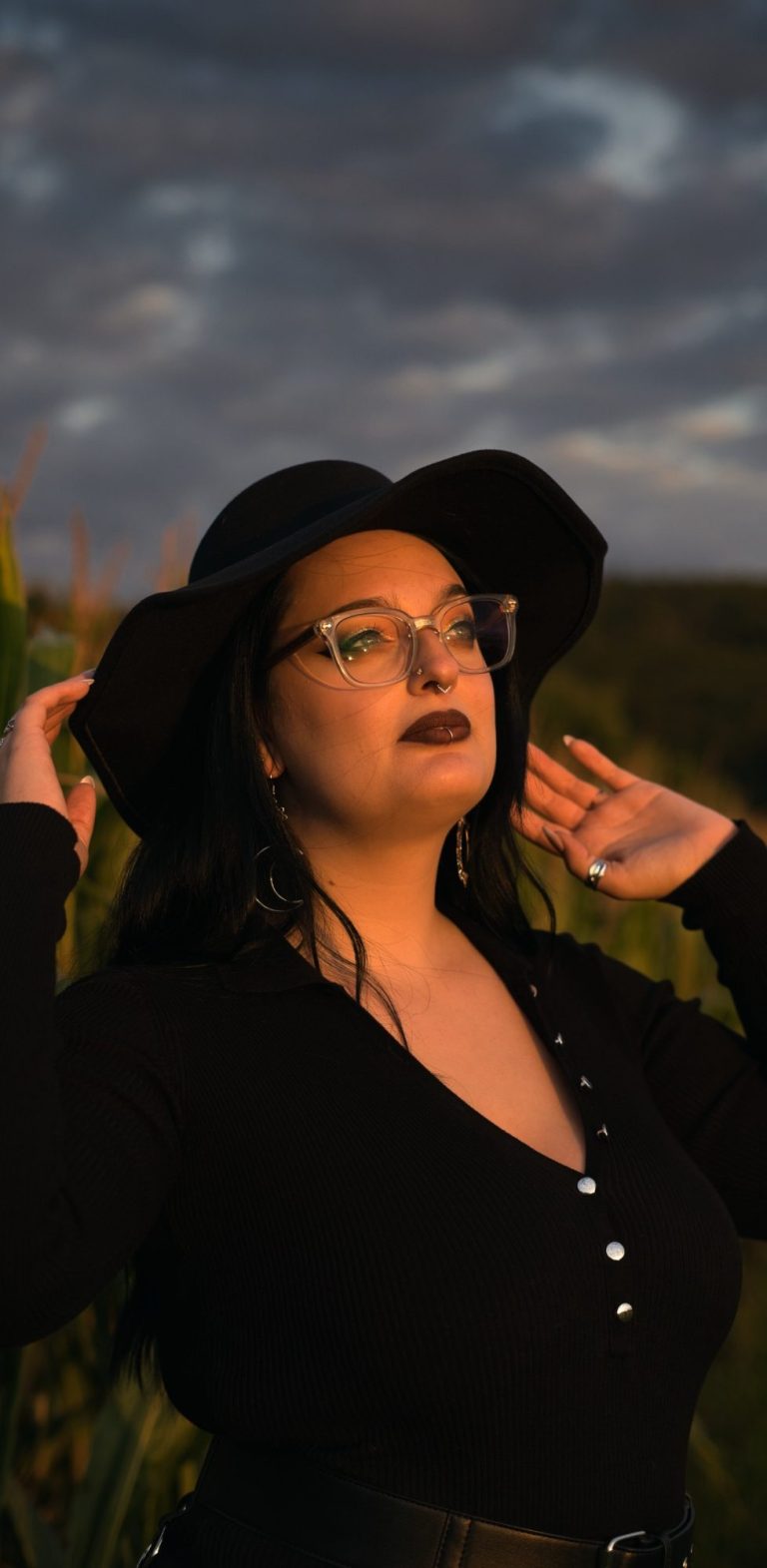 Frau mit langen, braunen Haaren in einem schwarzen Kleid, sitzt in einer bewaldeten Umgebung.