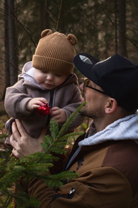Ein Vater hält ein Kleinkind im Wald, beide lächeln und der Junge spielt mit einem roten Gegenstand.