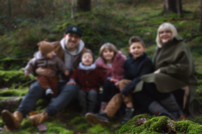 Familie sitzt auf Moos im Wald, umgeben von Bäumen, mit fröhlichen Gesichtern.