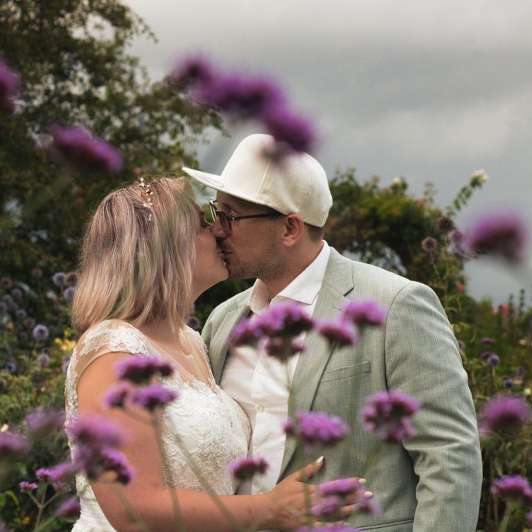 Ein Paar küsst sich umgeben von lila Blumen in einer romantischen Gartenumgebung.