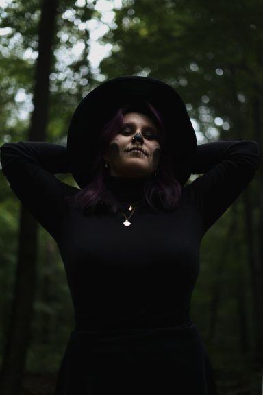 Frau mit lilanen Haaren und Hut, steht in einem dunklen Wald.