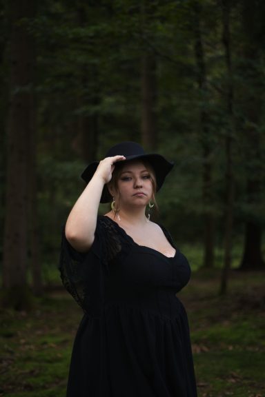 Frau in schwarzem Kleid und Hut steht im Wald, umgeben von Bäumen.