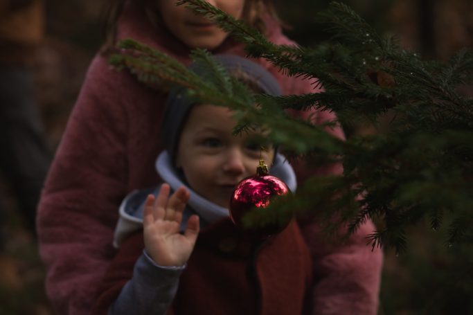 Ein Kind mit einer Weihnachtskugel in der Hand winkt, während es von hinten umarmt wird.