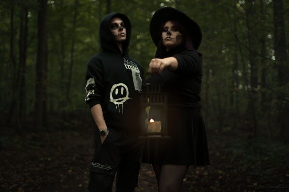 Zwei Personen im Wald, eine hält eine Laterne, beide tragen dunkle Kleidung.