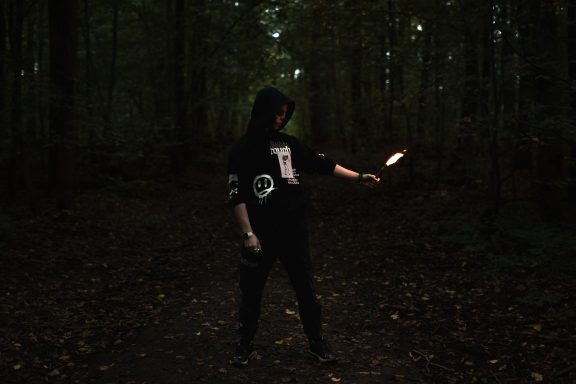 Person in dunkler Kleidung steht im Wald und hält eine brennende Fackel.